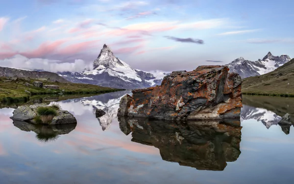 4K Ultra HD view of the Swiss Alps in Zermatt, showcasing a serene mountain landscape with a clear reflective lake and rugged rocks under a pastel sky.