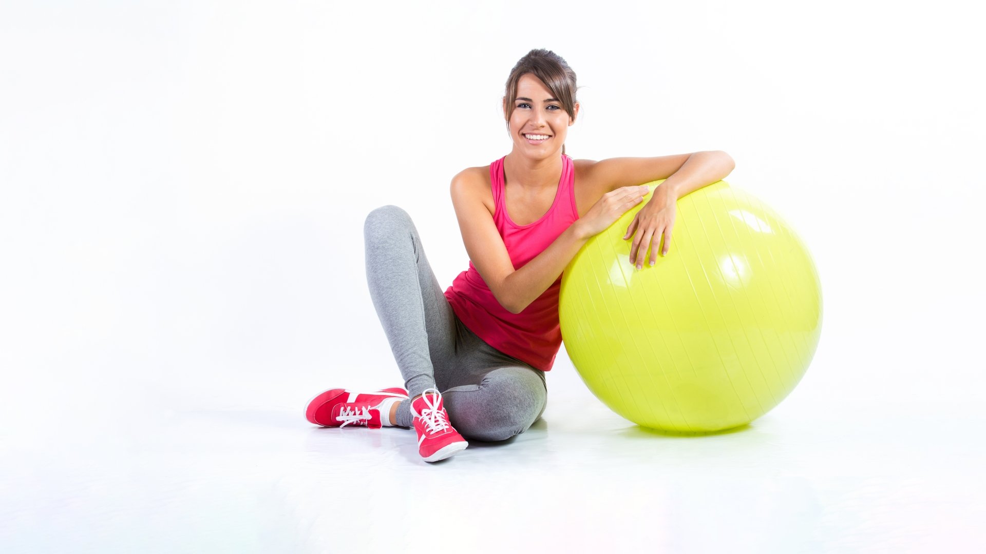 Brunette woman in fitness wear smiles while sitting on the floor, leaning on a large yellow exercise ball, captured in a bright 8K Ultra HD sports-themed desktop wallpaper.