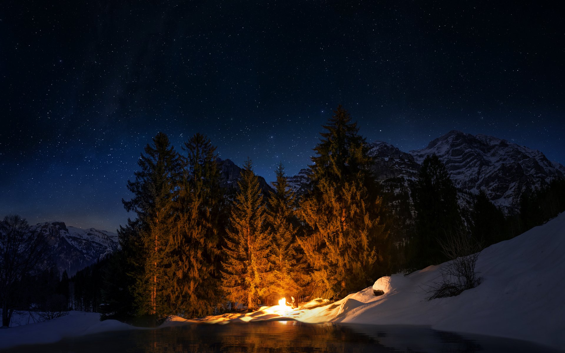 A bright bonfire illuminates a snowy winter forest under a starry night sky, captured in an HD nature photography desktop wallpaper.