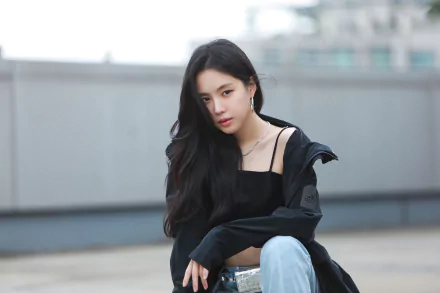 Female singer with long black hair seated on a rooftop in a casual jacket and crop top, moody urban 4K desktop wallpaper evoking music and style.