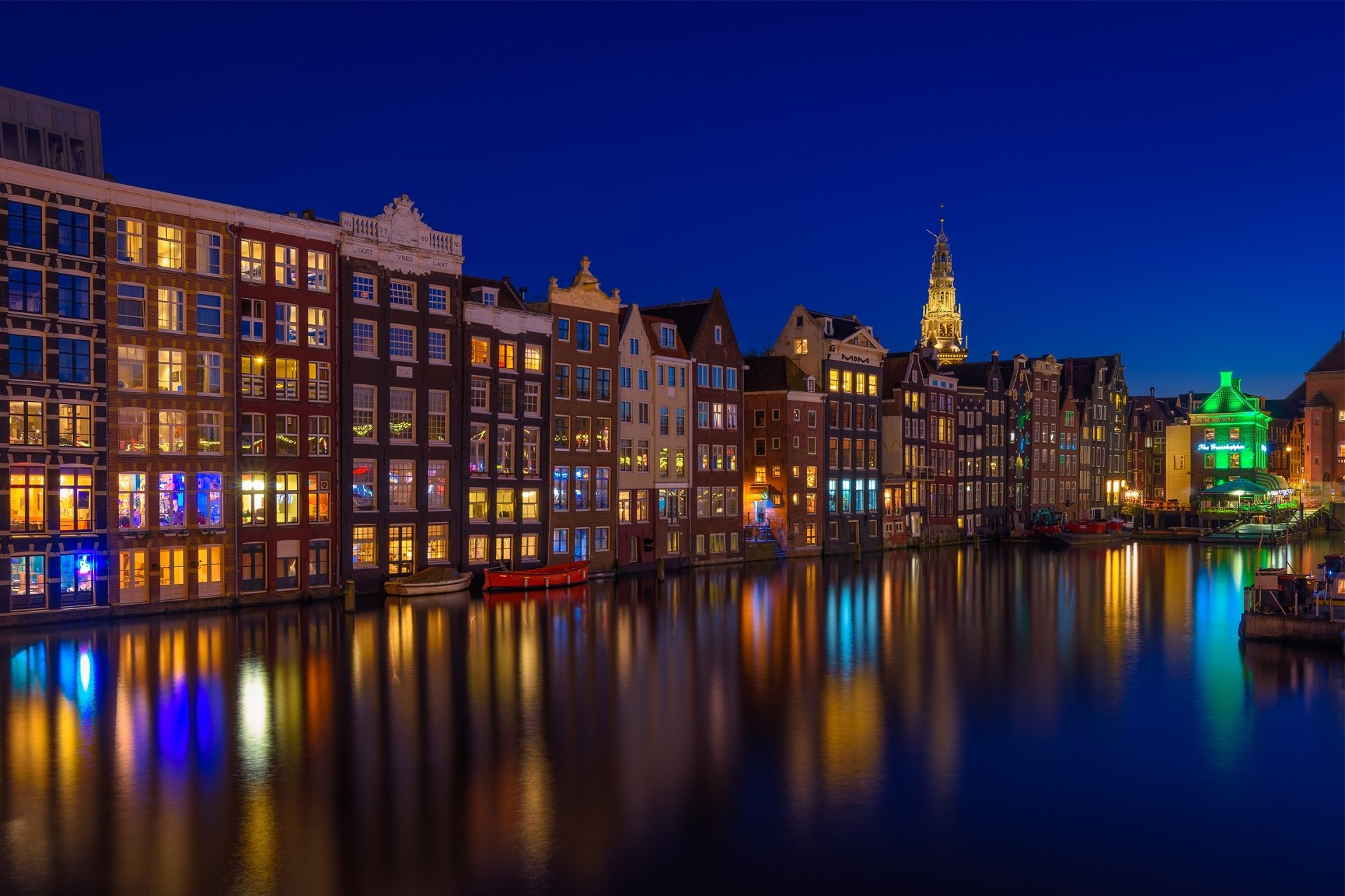 Night view of illuminated historic houses along a calm canal in Amsterdam, Netherlands, showcasing classic man-made architecture in an HD desktop wallpaper.