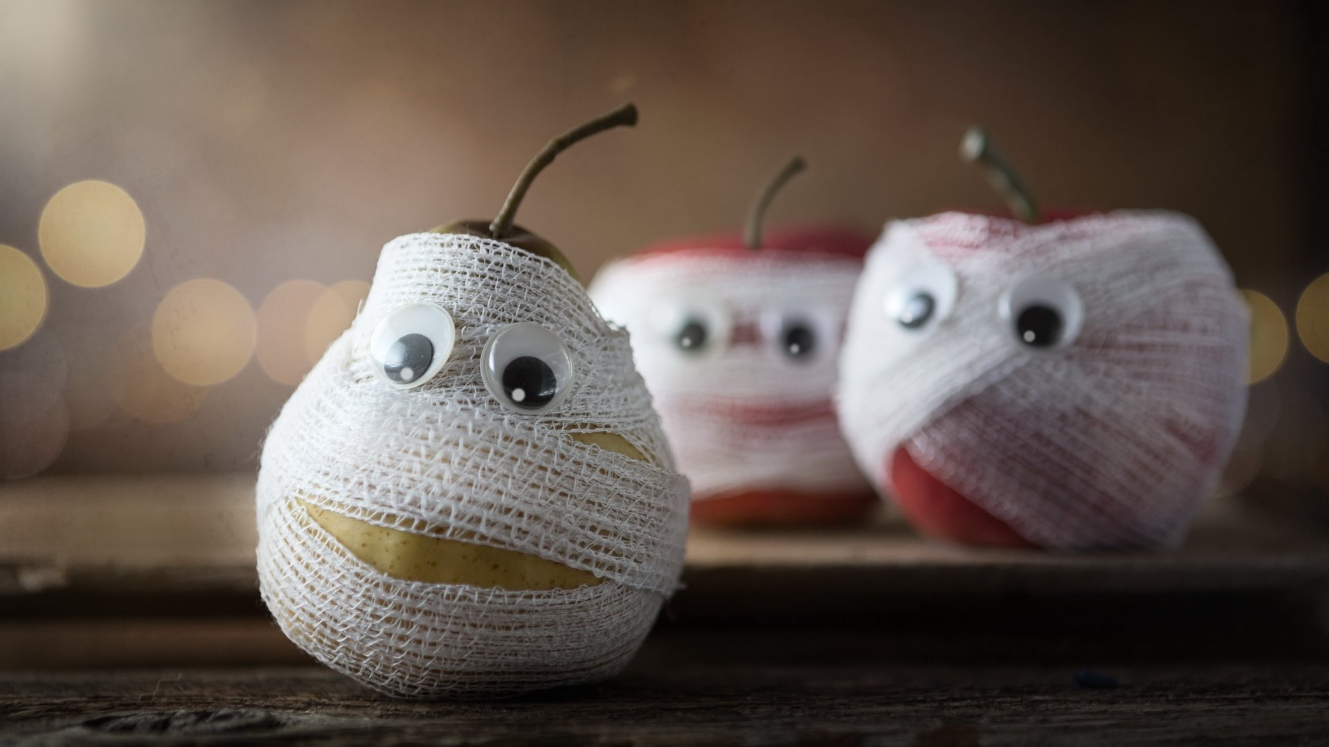 4K Ultra HD PC desktop wallpaper/background: funny food scene of a pear and two apples wrapped as mummies with googly eyes, staged on a rustic wooden tabletop.