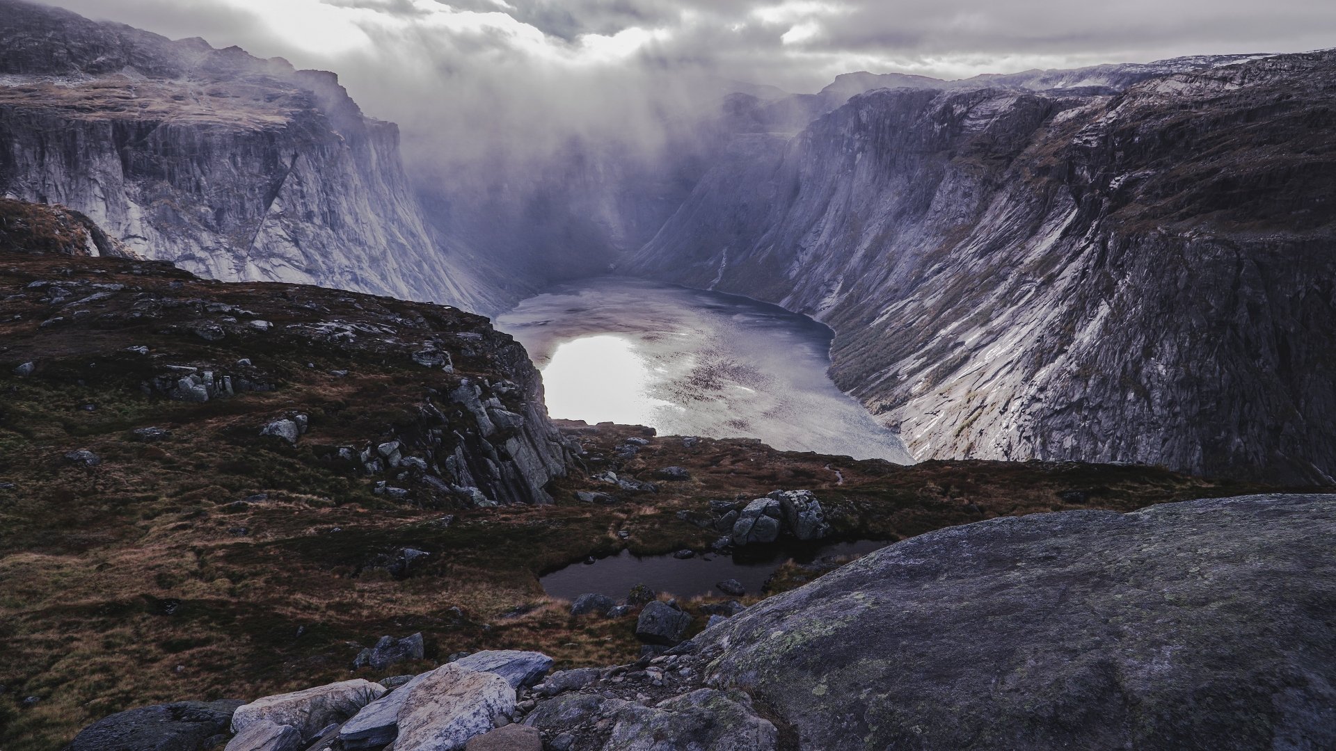 4K Ultra HD PC desktop wallpaper: dramatic mountain fjord framing a glassy lake below, rugged rock foreground and mist-filled valley under a moody sky.