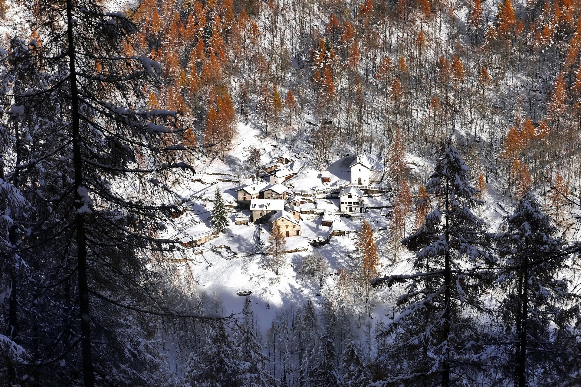 Snow-covered village nestled in a winter forest in Lombardy, Italy, surrounded by tall trees and autumn-colored foliage, captured in an HD desktop wallpaper.
