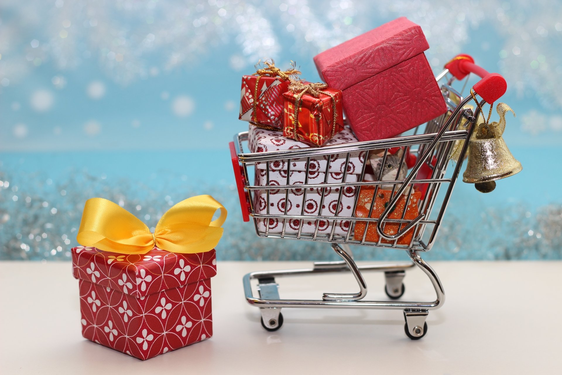 A festive 4K Ultra HD desktop wallpaper featuring a shopping cart filled with wrapped Christmas gifts and a bell, with a large gift box beside it against a snowy background.