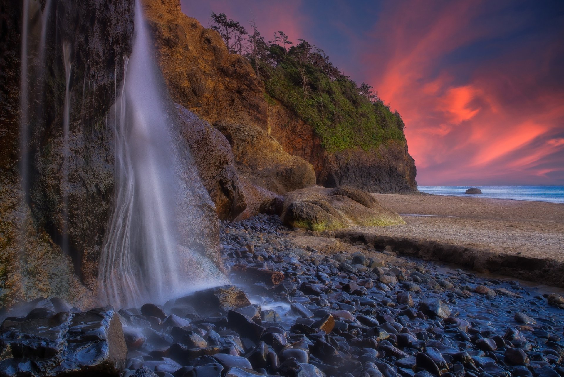 HD PC desktop wallpaper of a waterfall tumbling onto a stony Oregon coast at sunset, the Pacific Ocean framed by a vivid colorful sky.