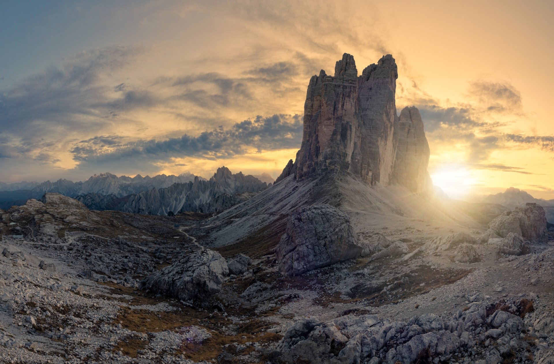Tre Cime di Lavaredo in the Dolomites at dawn, golden sun breaking behind jagged peaks over a rocky foreground — 4K Ultra HD PC desktop wallpaper and nature background.