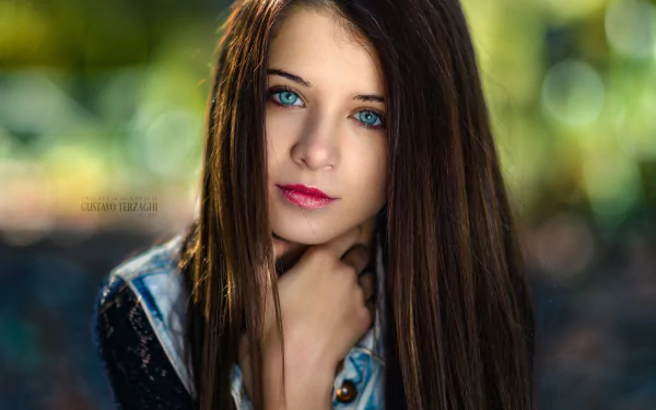 HD desktop wallpaper featuring a close-up portrait of brunette model Ariadna Velazquez with striking blue eyes and vibrant lipstick against a blurred natural background.