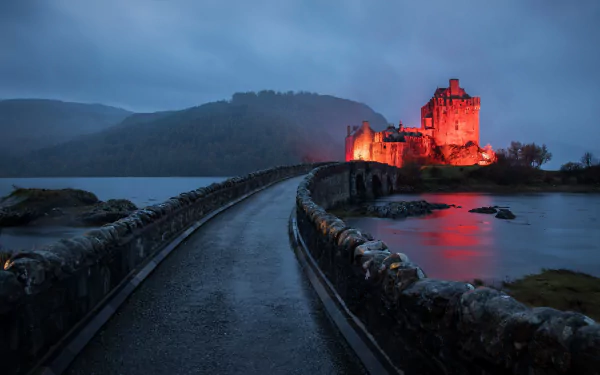 Eilean Donan Castle in Scotland illuminated at dusk, with a stone bridge leading across calm water under a moody sky, captured in 4K Ultra HD.