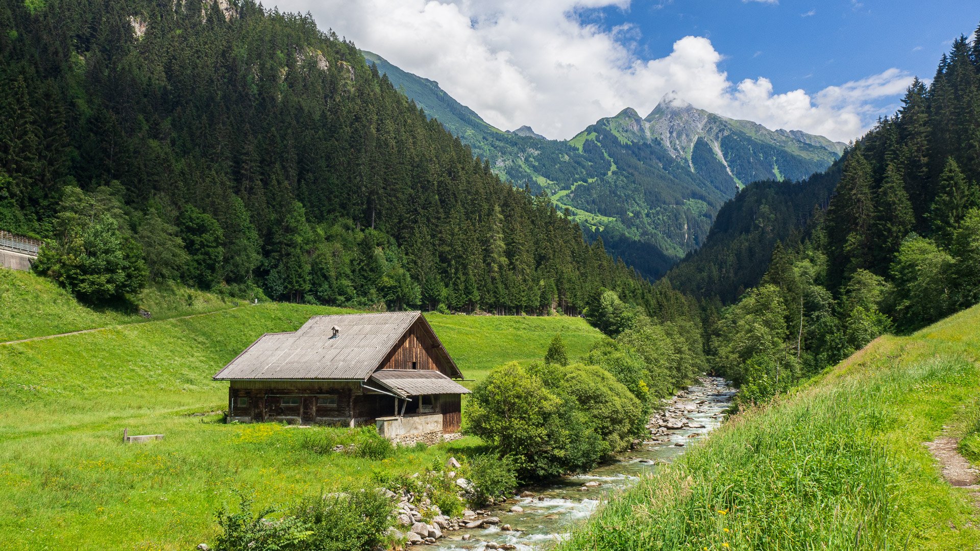 HD desktop wallpaper showcasing a peaceful alpine valley with a rustic cabin, flowing stream, dense forest, and towering Alps under a bright, partly cloudy sky.