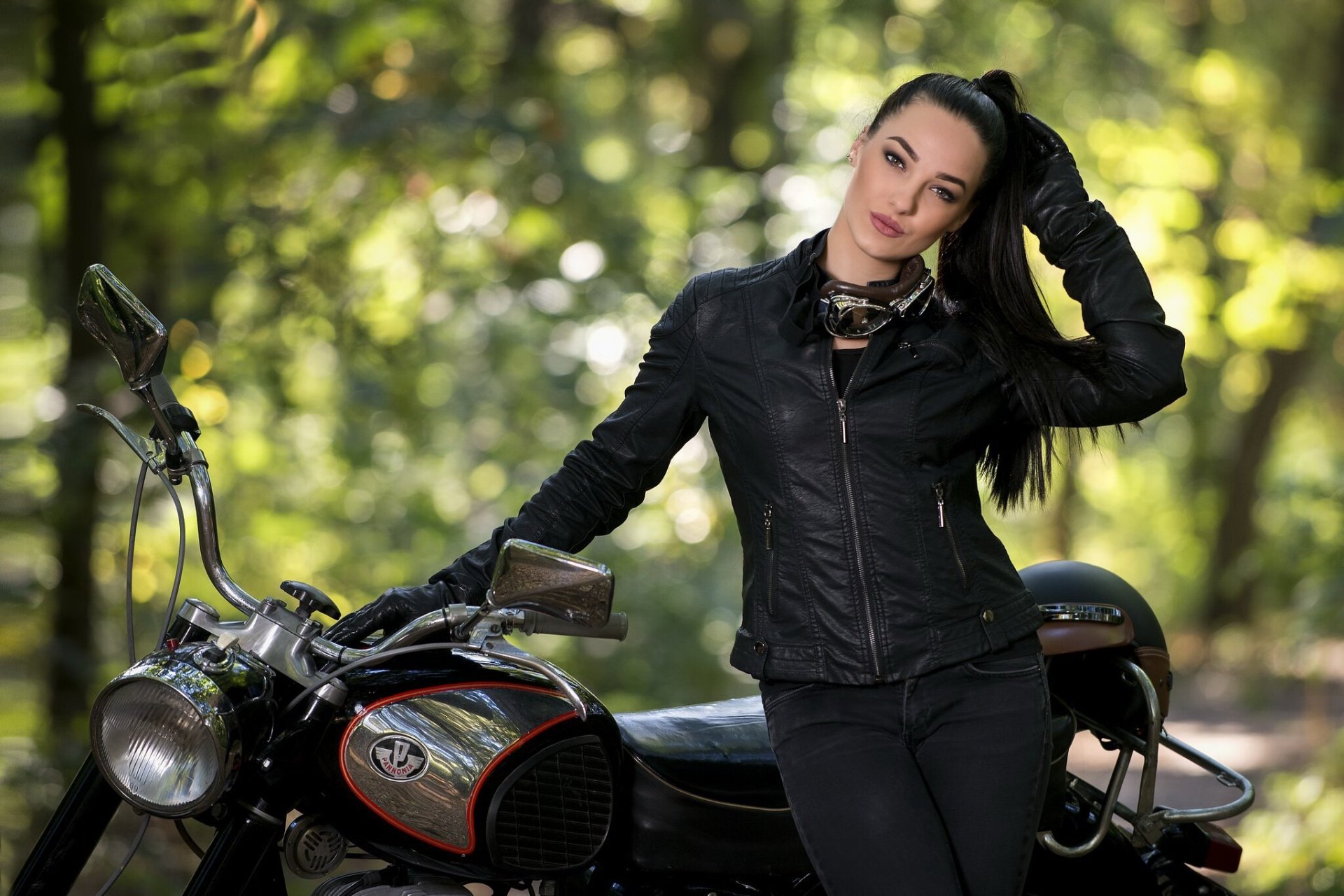 A woman with black hair in a ponytail wearing a black leather jacket leans on a Pannonia motorcycle, set against a blurred green natural background.