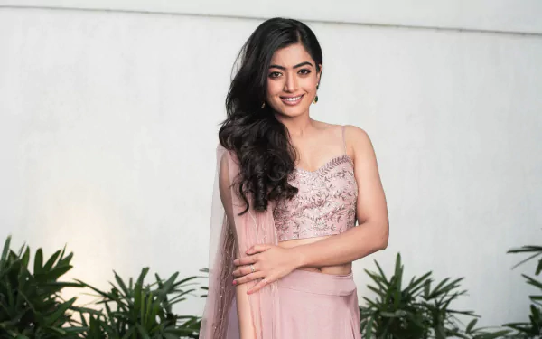 HD desktop wallpaper of a smiling woman in a pink dress, standing with folded arms against a leafy backdrop.