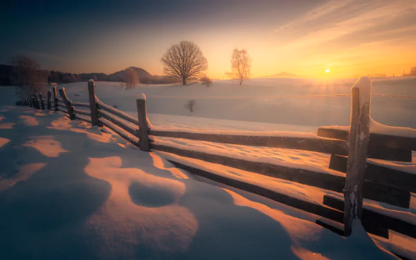 Snow-covered wooden fence stretches through a serene winter landscape at sunset, with bare trees and soft light creating a peaceful outdoor scene, captured in HD photography.