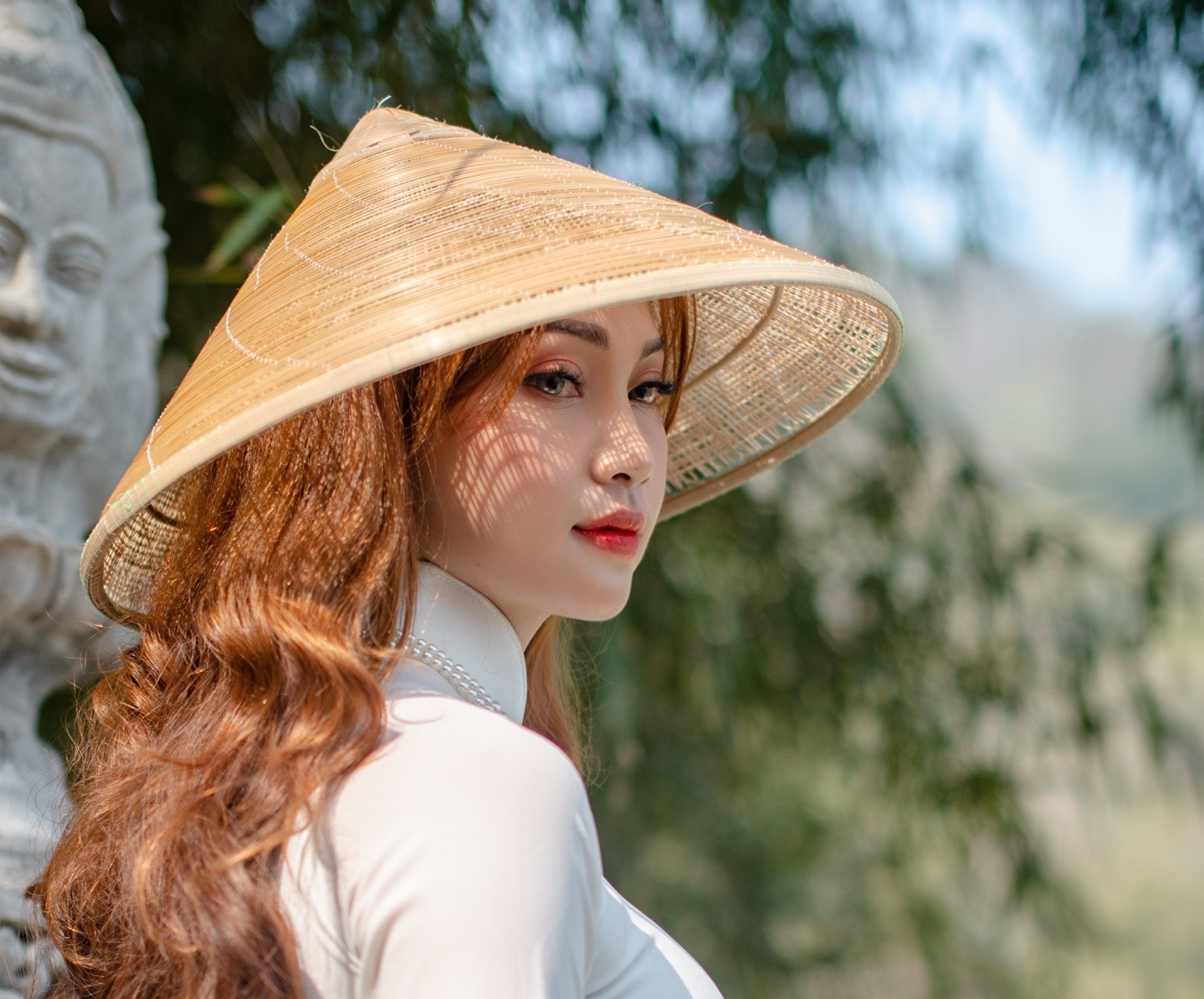 HD desktop wallpaper of a brunette Asian woman wearing a traditional conical hat, posed outdoors with soft natural lighting and a serene background.