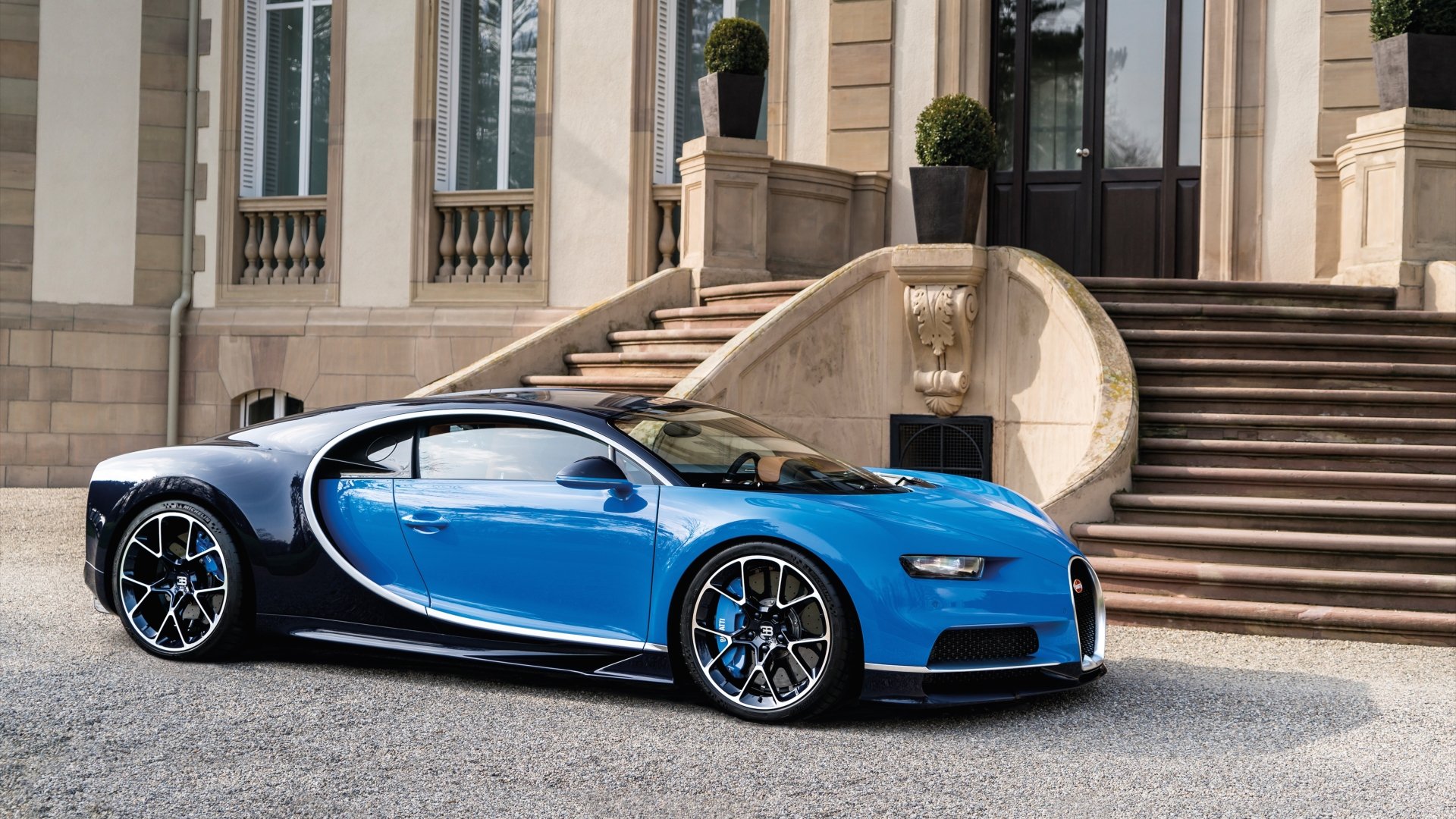 HD desktop wallpaper featuring a sleek blue Bugatti Chiron supercar parked in front of an elegant building with grand stairs and potted plants.