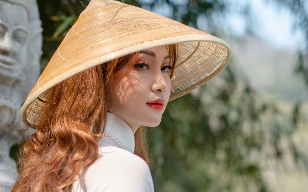 HD desktop wallpaper of a brunette Asian woman wearing a traditional conical hat, posed outdoors with soft natural lighting and a serene background.