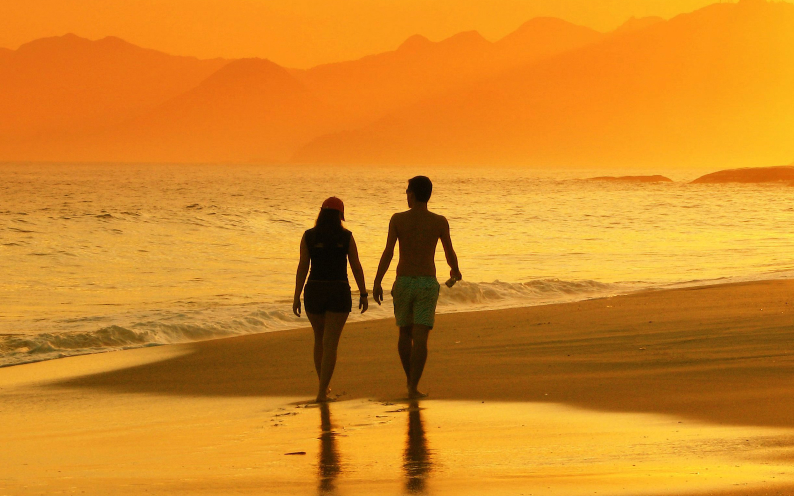 Sunset beach couple walking hand in hand along the orange ocean shore.