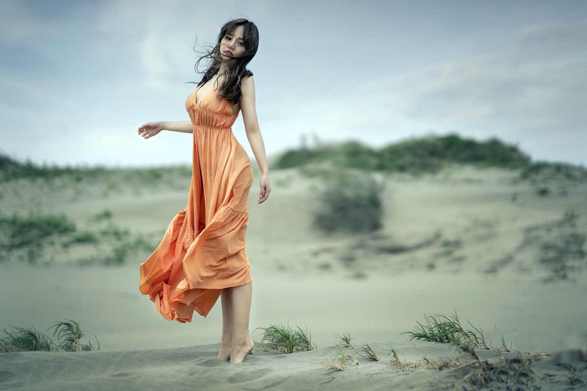 Brunette woman in a flowing orange dress — a model posing barefoot on windswept sand dunes, soft depth-of-field bokeh, HD desktop wallpaper background.