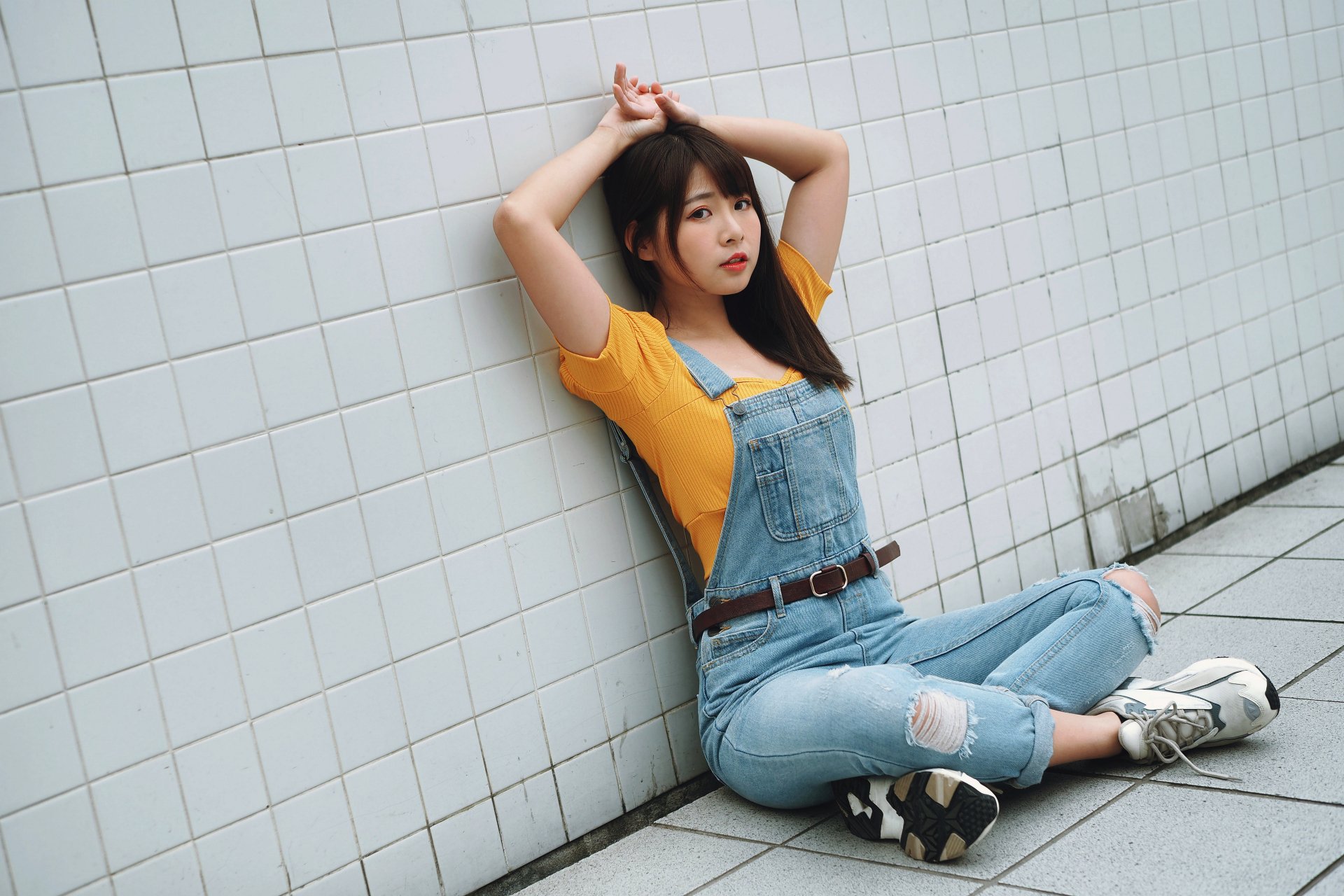 Asian brunette model wearing denim overalls and a yellow top sits against a white tiled wall in this 4K Ultra HD desktop wallpaper and background.