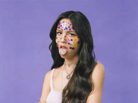 HD desktop wallpaper of American brunette singer and actress Olivia Rodrigo with colorful stickers on her face against a purple background.
