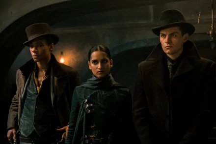 HD wallpaper of Shadow and Bone characters with Inej Ghafa in the center flanked by Jesper Fahey on the left and Kaz Brekker on the right, posing dramatically in a dimly lit setting.