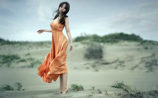 Brunette woman in a flowing orange dress — a model posing barefoot on windswept sand dunes, soft depth-of-field bokeh, HD desktop wallpaper background.
