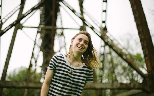 HD desktop wallpaper featuring a smiling person in a striped shirt standing in front of a bridge structure, associated with Julien Baker.