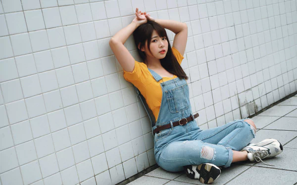 Asian brunette model wearing denim overalls and a yellow top sits against a white tiled wall in this 4K Ultra HD desktop wallpaper and background.