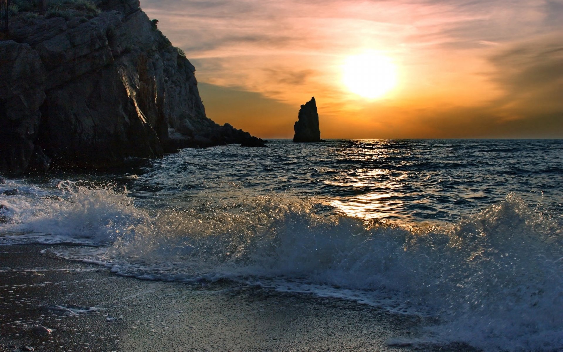 A stunning HD wallpaper showcasing a sunset over the ocean, with waves gently lapping at the shore and a silhouette of a rock formation against the horizon. Nature's beauty captured in vivid detail.