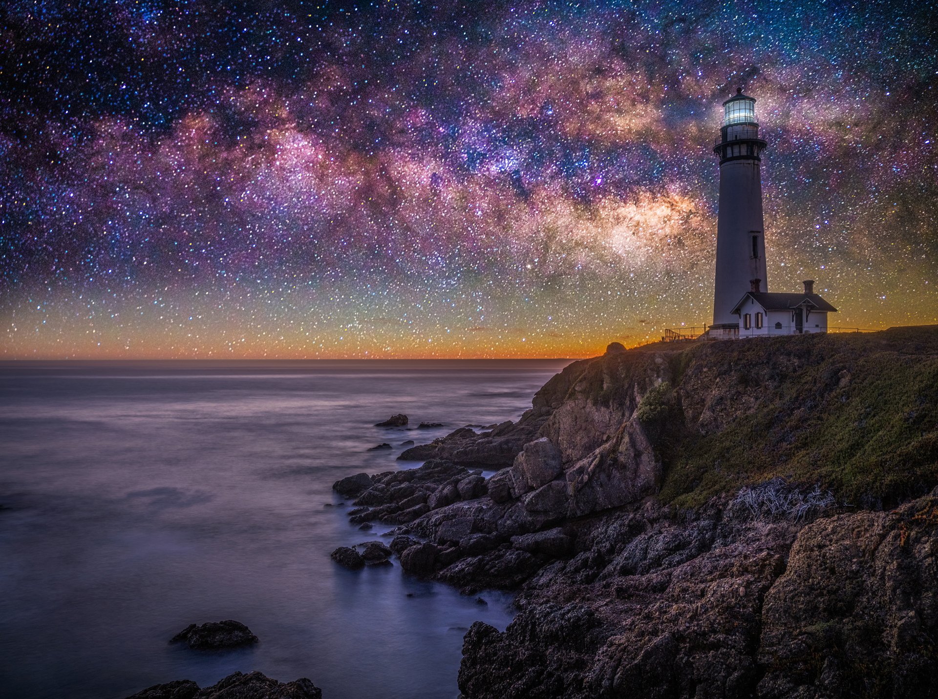 4K Ultra HD PC desktop background: a man-made lighthouse atop rocky cliffs at night beneath a vivid starry sky and Milky Way, waves lapping the coast.