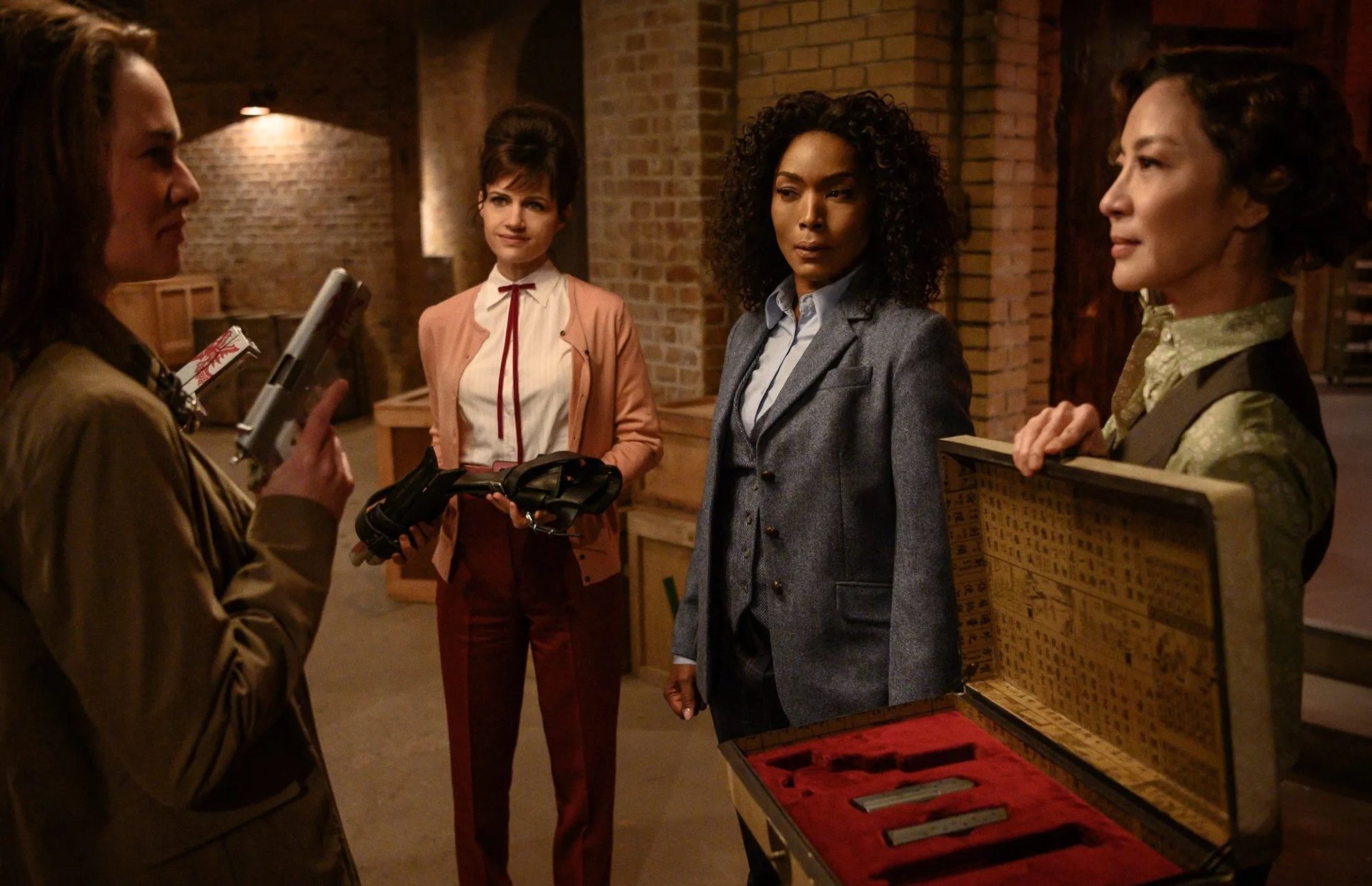 Four women in tailored outfits stand in a dim brick room exchanging guns and a wooden case of knives — cinematic action-movie promotional HD wallpaper.