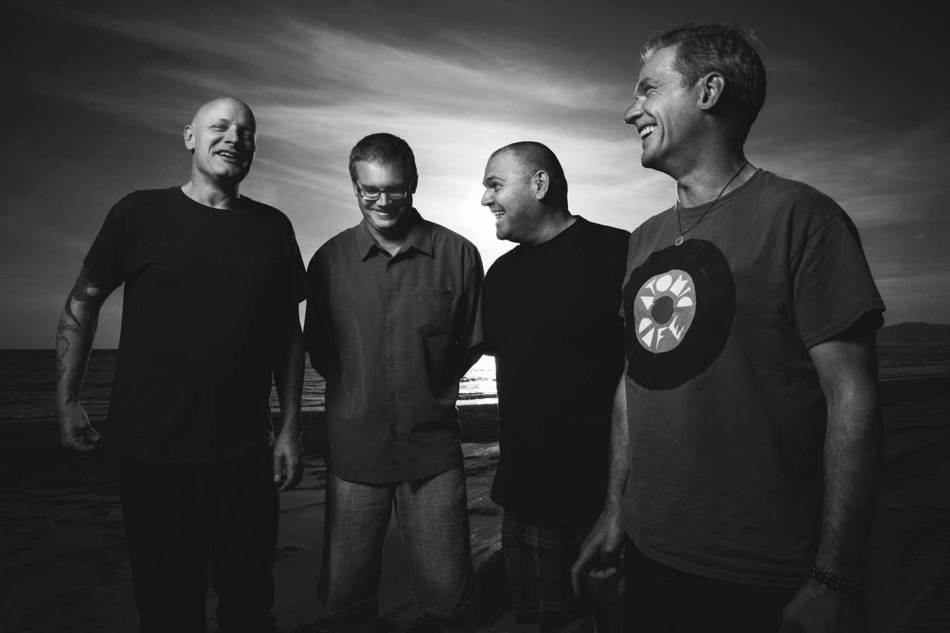 Black and white HD wallpaper featuring four smiling men standing together on a beach at sunset, evoking themes associated with the Descendents.