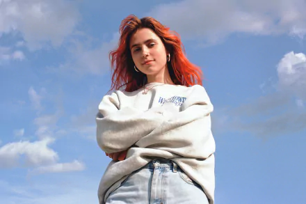 HD desktop wallpaper featuring a person with red hair in a casual grey sweatshirt and blue jeans against a sky background.