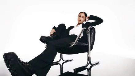 HD desktop wallpaper featuring American brunette actress and singer Olivia Rodrigo seated confidently on a modern black chair against a white background.