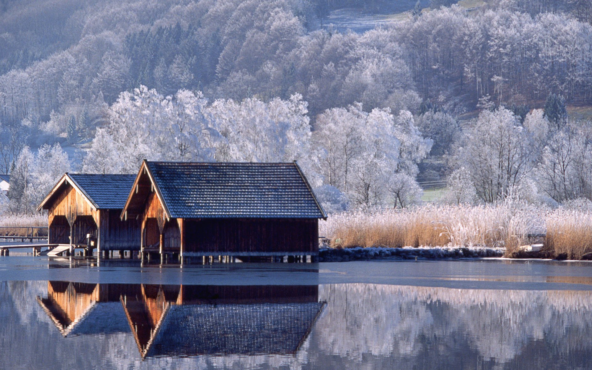 HD desktop wallpaper showing a serene winter landscape with wooden boathouses on a calm lake, surrounded by frost-covered trees in a natural setting.