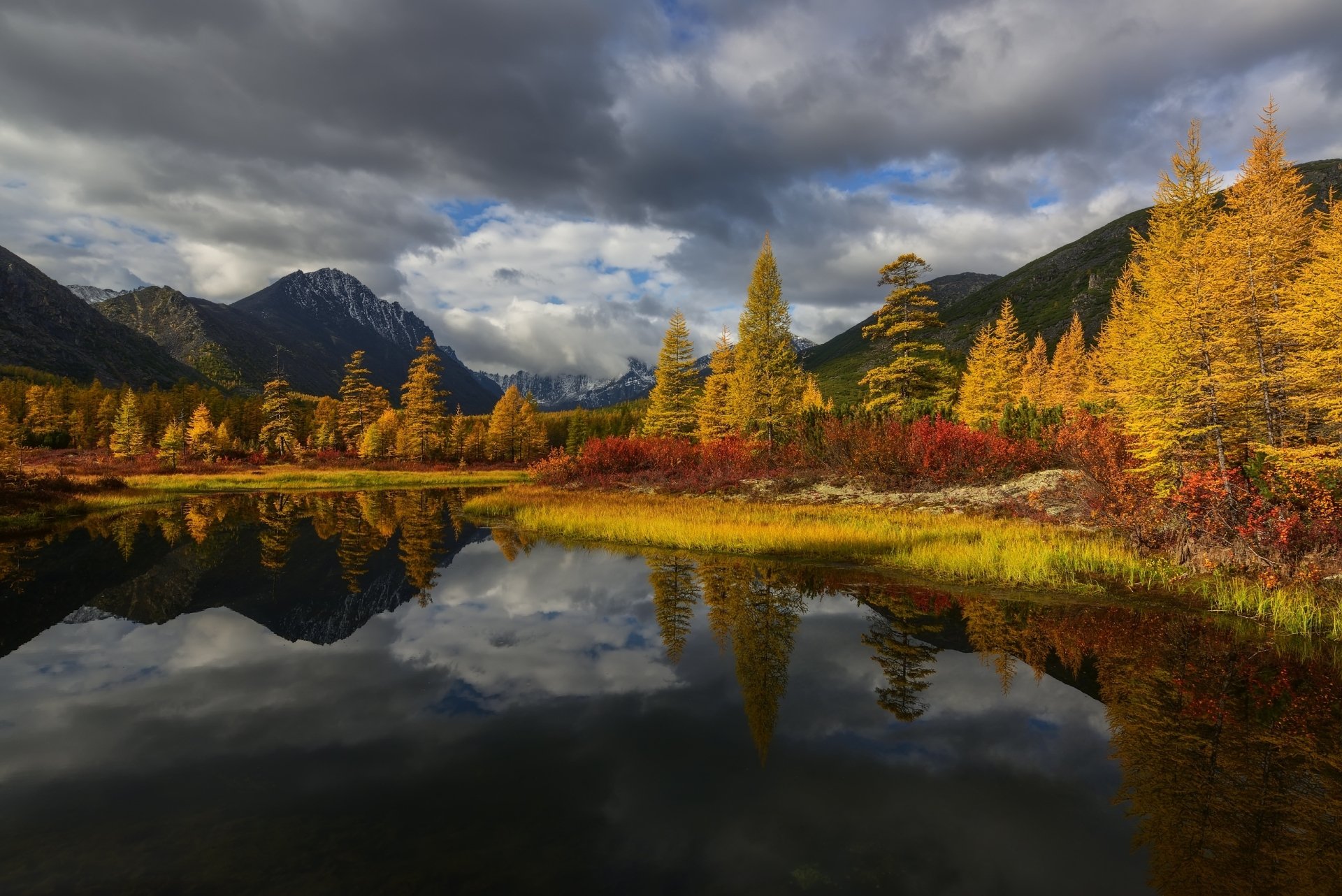 2K Quad HD PC desktop wallpaper/background: fall nature scene with golden larches and red shrubs reflected in a glassy mountain lake beneath dramatic cloudy skies.