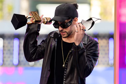 HD PC wallpaper of a rapper and music artist onstage wearing a black cap, sunglasses and leather jacket, holding a trophy, with colorful blurred stage lights in the background.