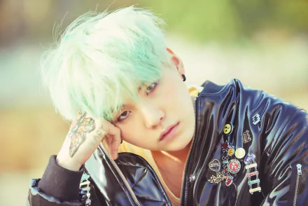HD PC desktop wallpaper of a mint‑haired K-pop singer in a studded leather jacket with pins and tattoos, gazing pensively — music-themed portrait.