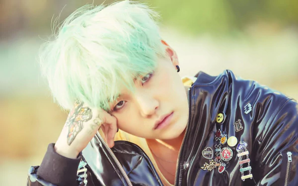 HD PC desktop wallpaper of a mint‑haired K-pop singer in a studded leather jacket with pins and tattoos, gazing pensively — music-themed portrait.