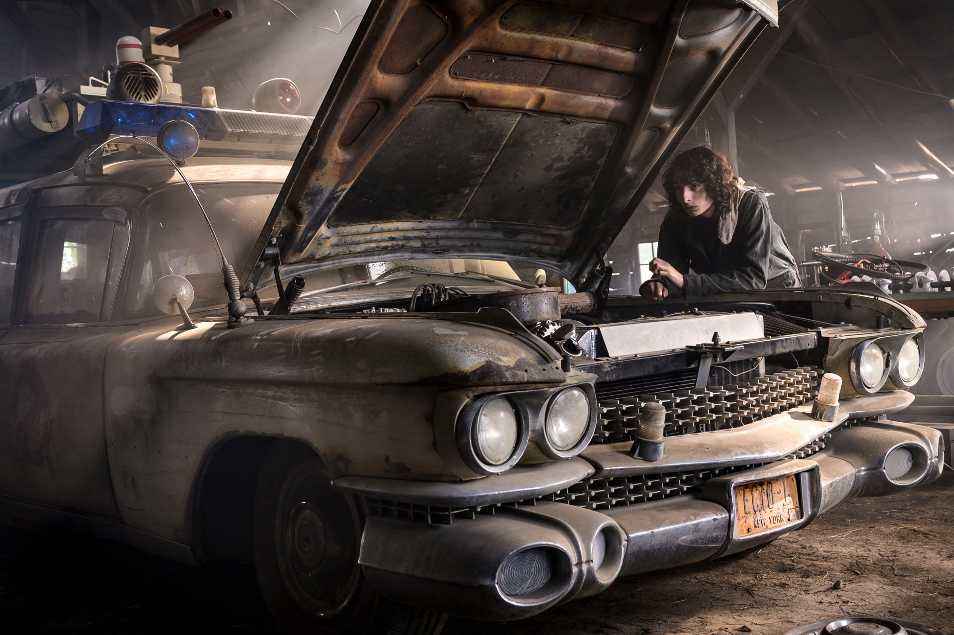 Finn Wolfhard in a scene from Ghostbusters: Afterlife, working under the hood of a vintage car in a dimly lit garage, captured in 4K Ultra HD for PC wallpaper.