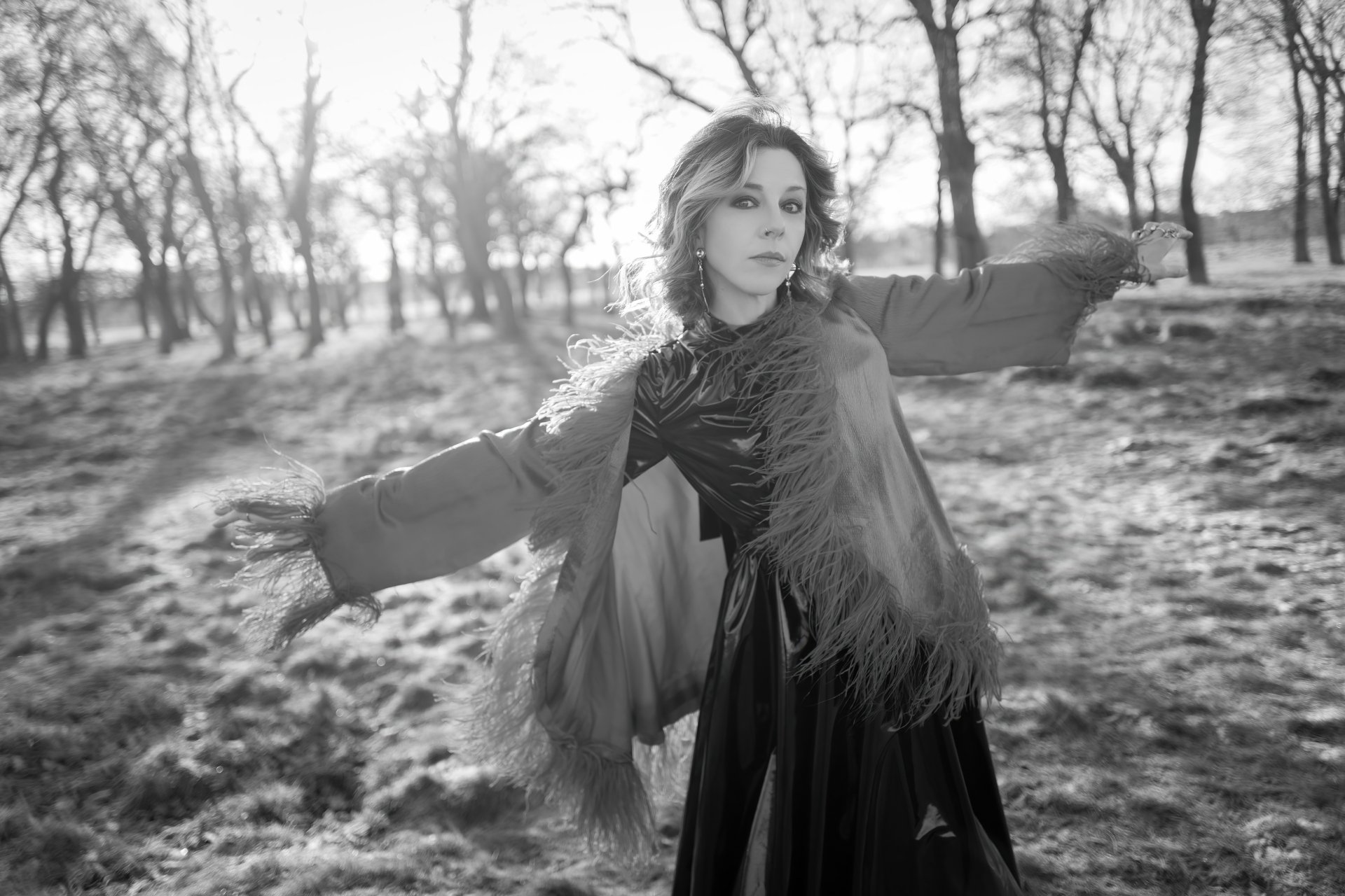Black-and-white 5K PC desktop background: woman in a flowing gown with feathered sleeves standing in a sunlit, leafless field, arms gently outstretched.