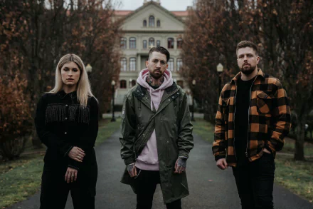 A moody HD desktop wallpaper featuring the band Spiritbox, with three members standing confidently in a park setting, surrounded by autumn trees and a historic building in the background.