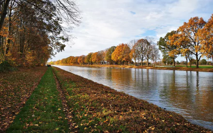 A serene HD PC desktop wallpaper of a river flanked by autumn trees and a grassy path under a partly cloudy sky, showcasing nature's vibrant fall colors.