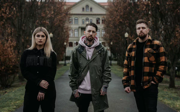 A moody HD desktop wallpaper featuring the band Spiritbox, with three members standing confidently in a park setting, surrounded by autumn trees and a historic building in the background.