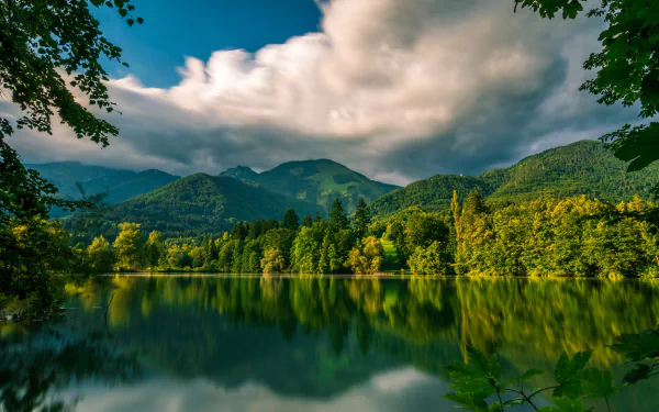 HD desktop wallpaper of Lake Črnava in Slovenia, featuring lush greenery, calm lake waters reflecting the cloudy sky, and surrounding forested hills under vibrant light.