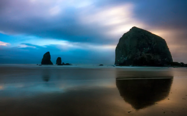 8K Ultra HD desktop wallpaper showcasing a serene beach scene with large rock formations and a calm, reflective shoreline under a dramatic, cloudy sky.
