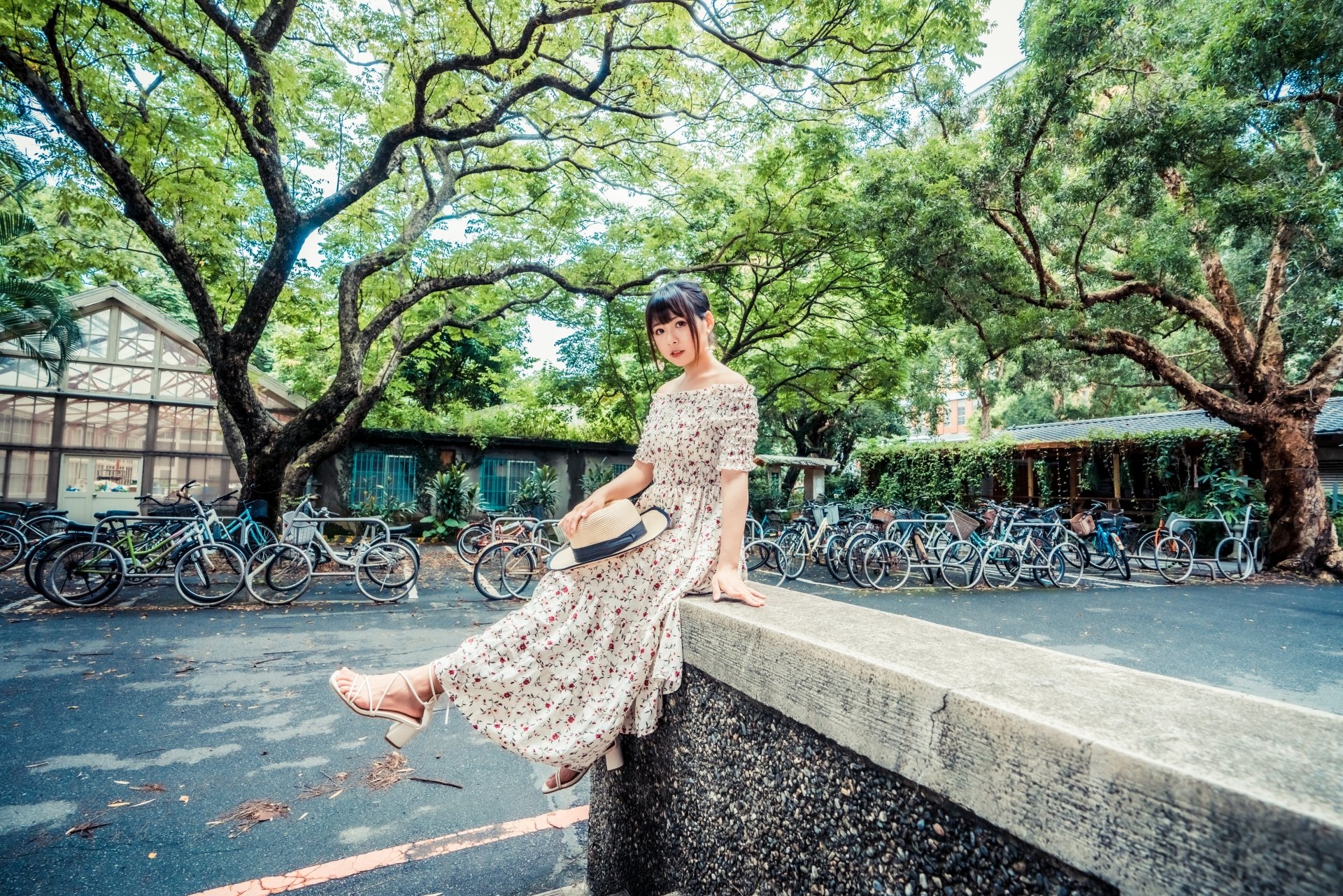 Asian woman in a floral dress poses outdoors by a stone ledge under large green trees, with bicycles parked in the background, captured in 4K Ultra HD detail.