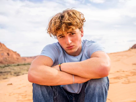 Benson Boone sitting with arms crossed in a desert landscape, featured in a 4K Ultra HD music-themed PC desktop wallpaper.