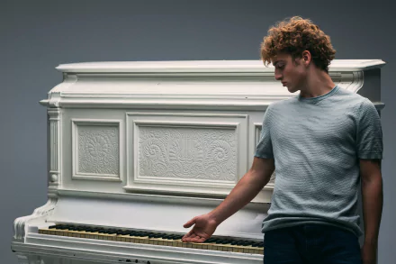 Benson Boone stands beside a white piano, looking down with a contemplative expression, captured in a 4K Ultra HD music-themed desktop wallpaper.