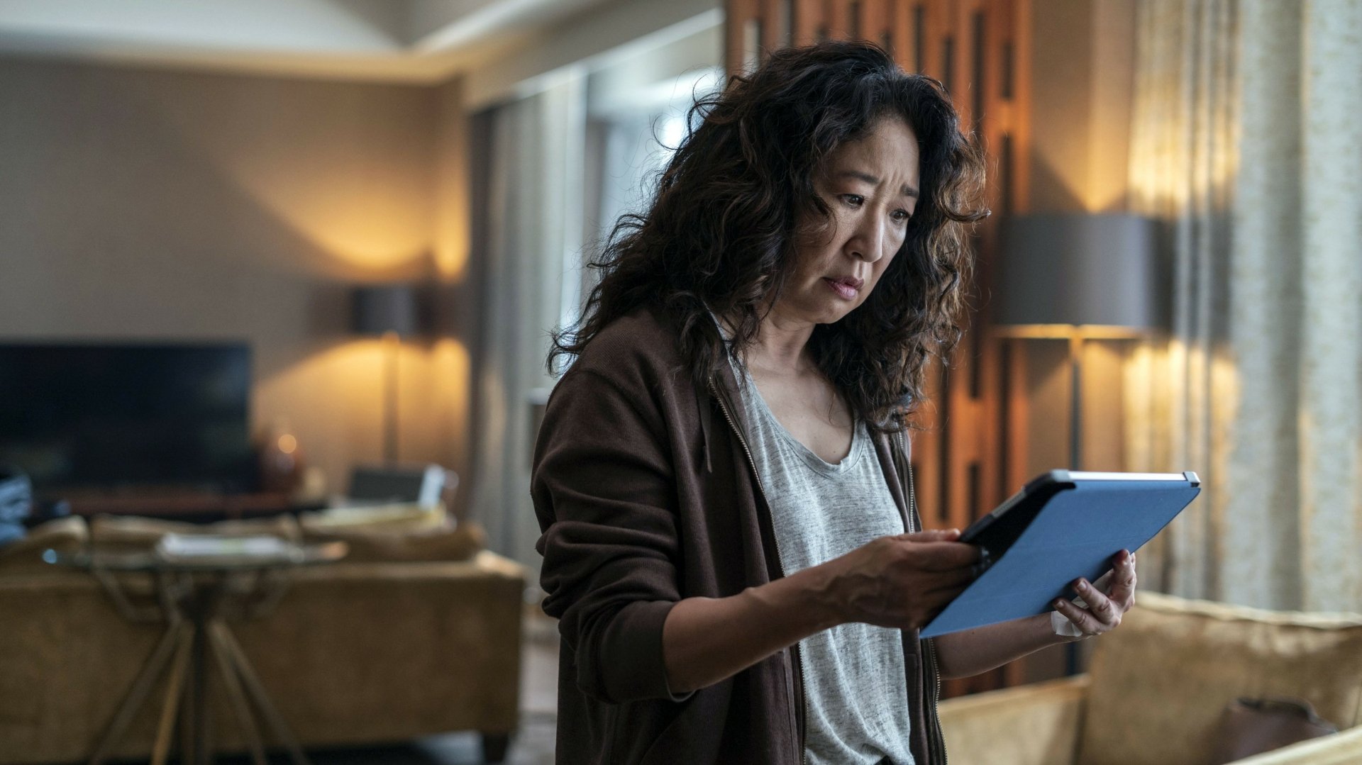 Killing Eve TV show 2K Quad HD PC desktop wallpaper: a woman in a dim living room studies a tablet with a tense expression, warm moody lighting and lived-in decor.
